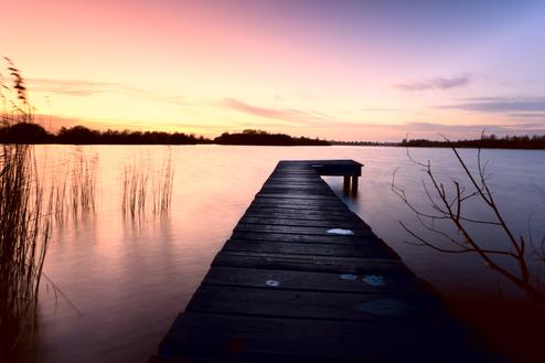 Holzsteg führt über ruhiges Wasser, umgeben von Gräsern und einem farbenprächtigen Sonnenuntergang.